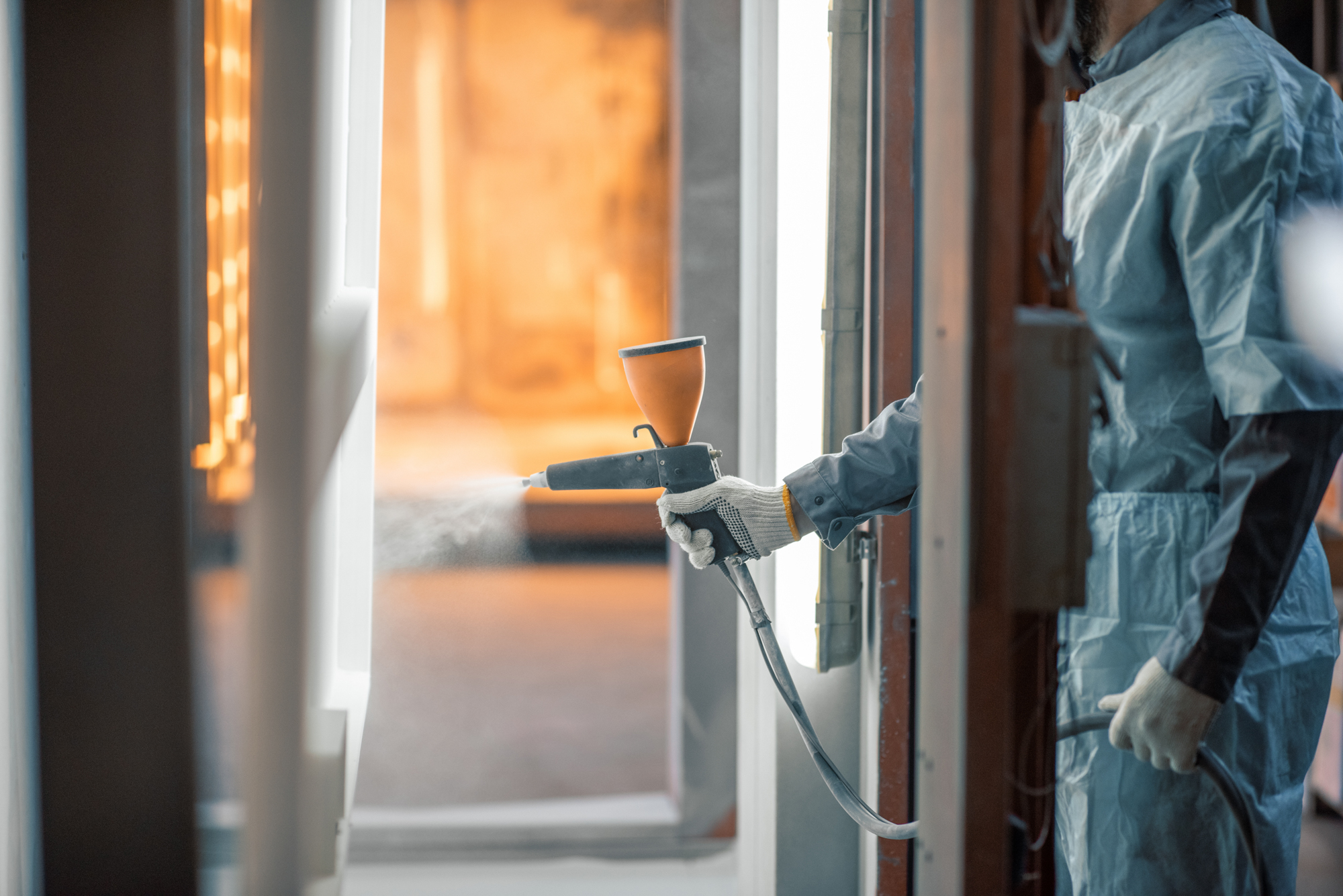 Työntekijä suorittaa jauhemaalausta teollisessa ympäristössä, käyttäen ruiskua suojavarustuksessa. Maalaussuutin tuottaa tasaisen jauhepinnoitesuihkun käsiteltävälle pinnalle.