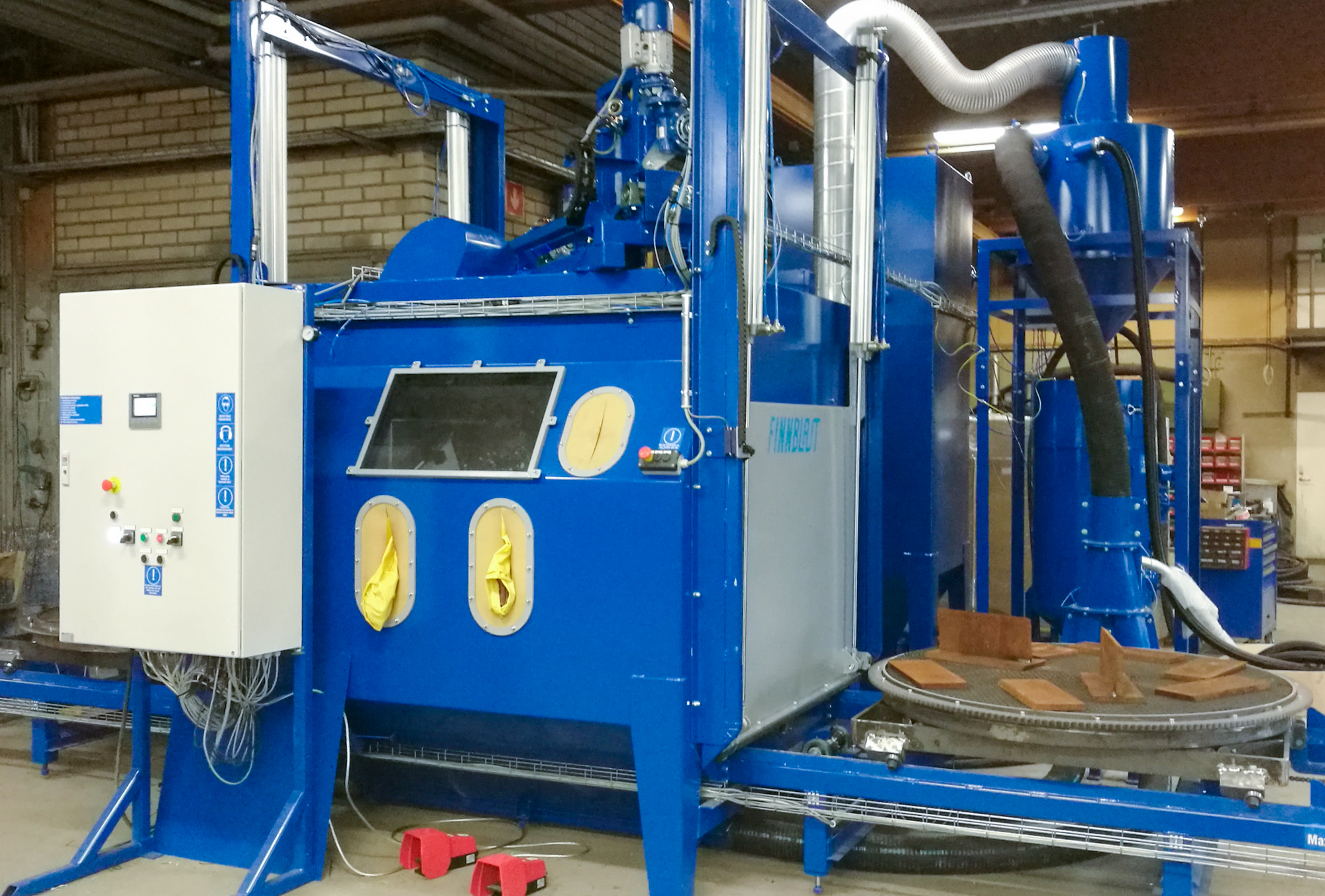 Automaattinen raepuhalluskone teollisuushallissa: sininen kaksipöytäinen laitteisto, jossa näkyy pyöreä työstöpöytä, ohjauspaneeli ja imurointi- sekä pölynpoistojärjestelmä teolliseen pintakäsittelyyn.