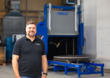 Jollmaxin toimitusjohtaja Markus Olshin seisoo teollisuushallissa hymyillen, takanaan sininen Finnblast-merkkinen raepuhalluskone.