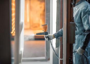 Työntekijä suorittaa jauhemaalausta teollisessa ympäristössä, käyttäen ruiskua suojavarustuksessa. Maalaussuutin tuottaa tasaisen jauhepinnoitesuihkun käsiteltävälle pinnalle.